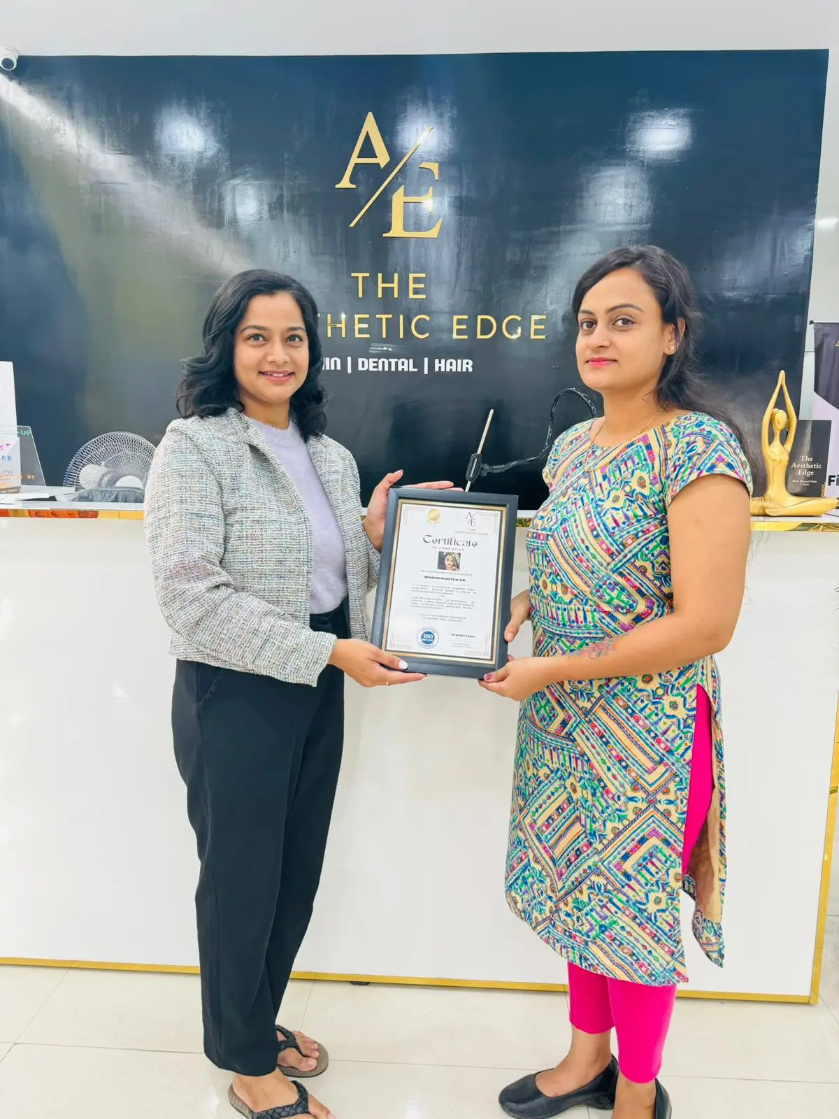 An academy student receiving her framed certificate of completion from a member of the team, in front of The Aesthetic Edge branded wall