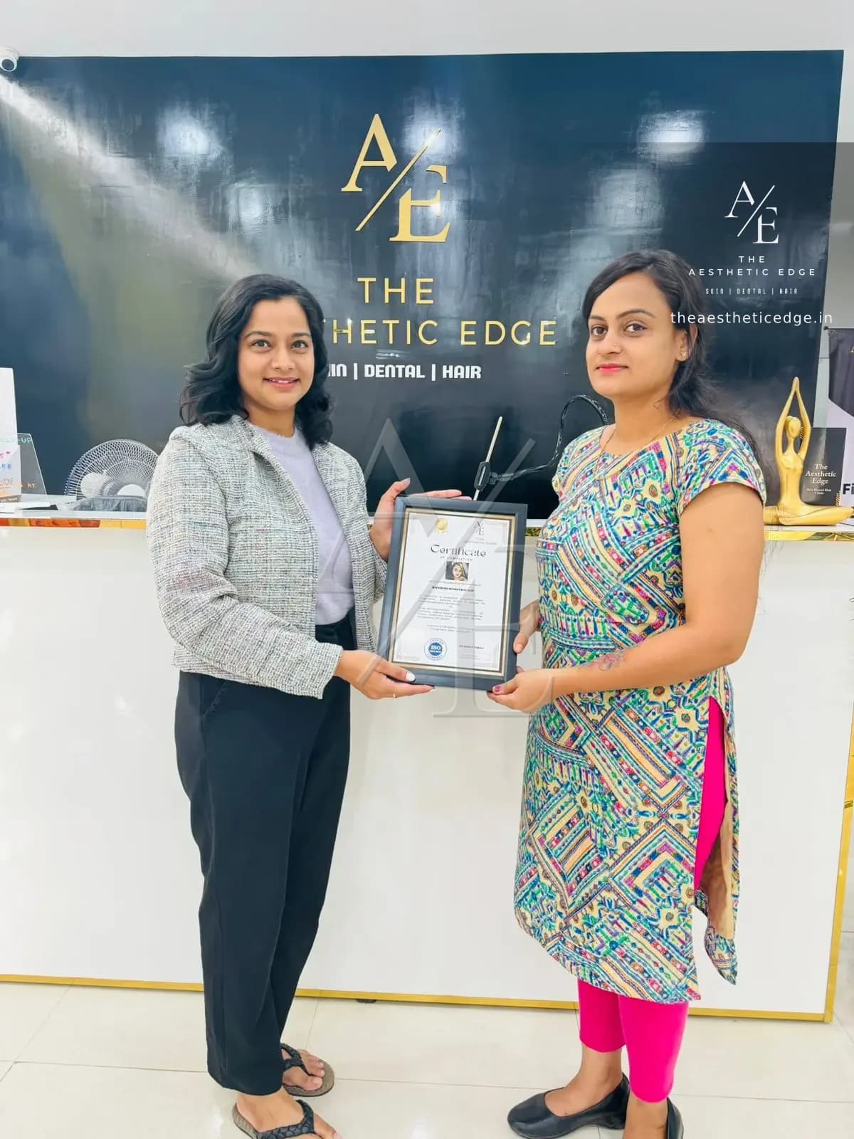 An academy student receiving her framed certificate of completion from a member of the team, in front of The Aesthetic Edge branded wall